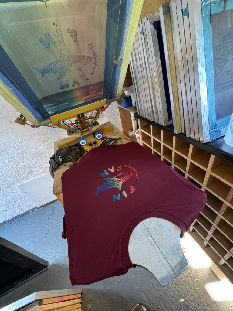 A burgundy Fin Fan T-shirt on the screen printing press, with the screen lifted above it